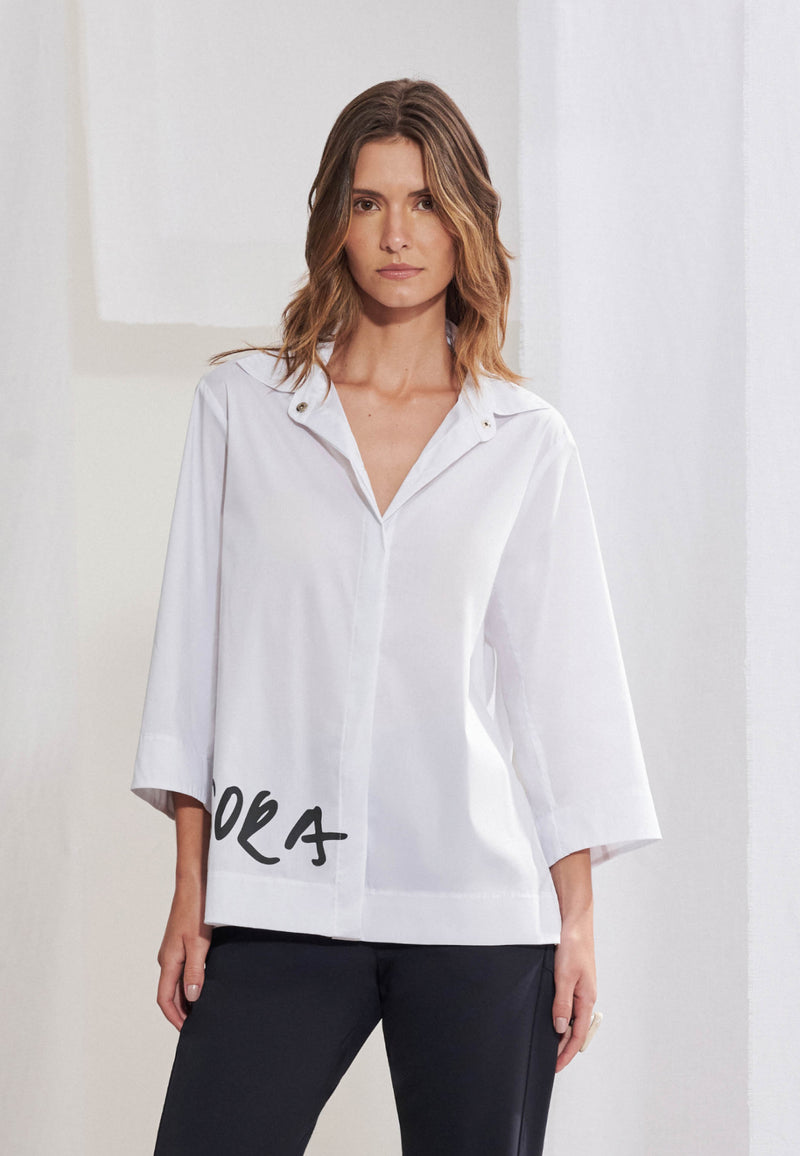 Mulher vestindo camisa oversized em tricoline macio com silhueta evasê. Gola colarinho, mangas 3/4 com punhos largos e fechamento frontal com botões ocultos. Na lateral inferior direita da barra, estampa tipográfica estilizada com a palavra "AGORA" em preto. Usada por fora de calça de alfaiataria escura.