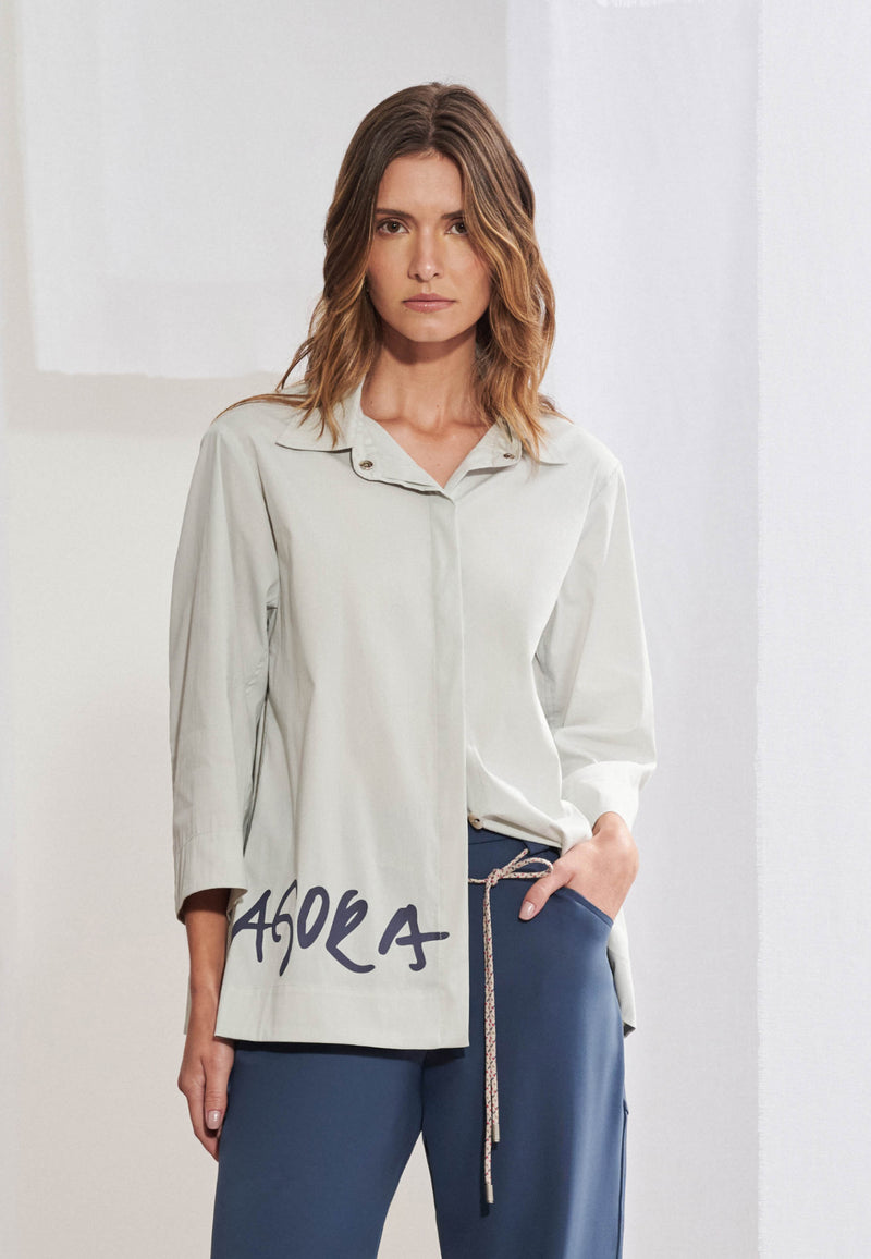 Mulher vestindo camisa oversized em tricoline macio com silhueta evasê. Gola colarinho, mangas 3/4 com punhos largos e fechamento frontal com botões ocultos. Na lateral inferior direita da barra, estampa tipográfica estilizada com a palavra "AGORA" em preto. Usada por fora de calça de alfaiataria escura.