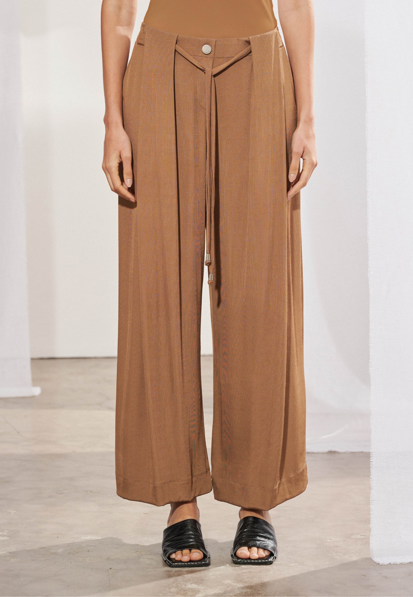 Calça pantalona longa em malha canelada, vista de frente. Modelagem ampla com pregas frontais profundas, cordão para amarração no cós com ponteiras metálicas e botão decorativo. Pernas largas com caimento fluido até o tornozelo.