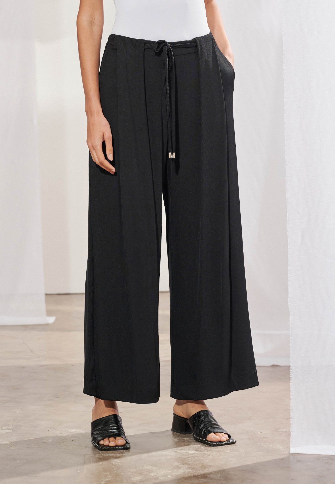 Calça pantalona longa em malha canelada, vista de frente. Modelagem ampla com pregas frontais profundas, cordão para amarração no cós com ponteiras metálicas e botão decorativo. Pernas largas com caimento fluido até o tornozelo.