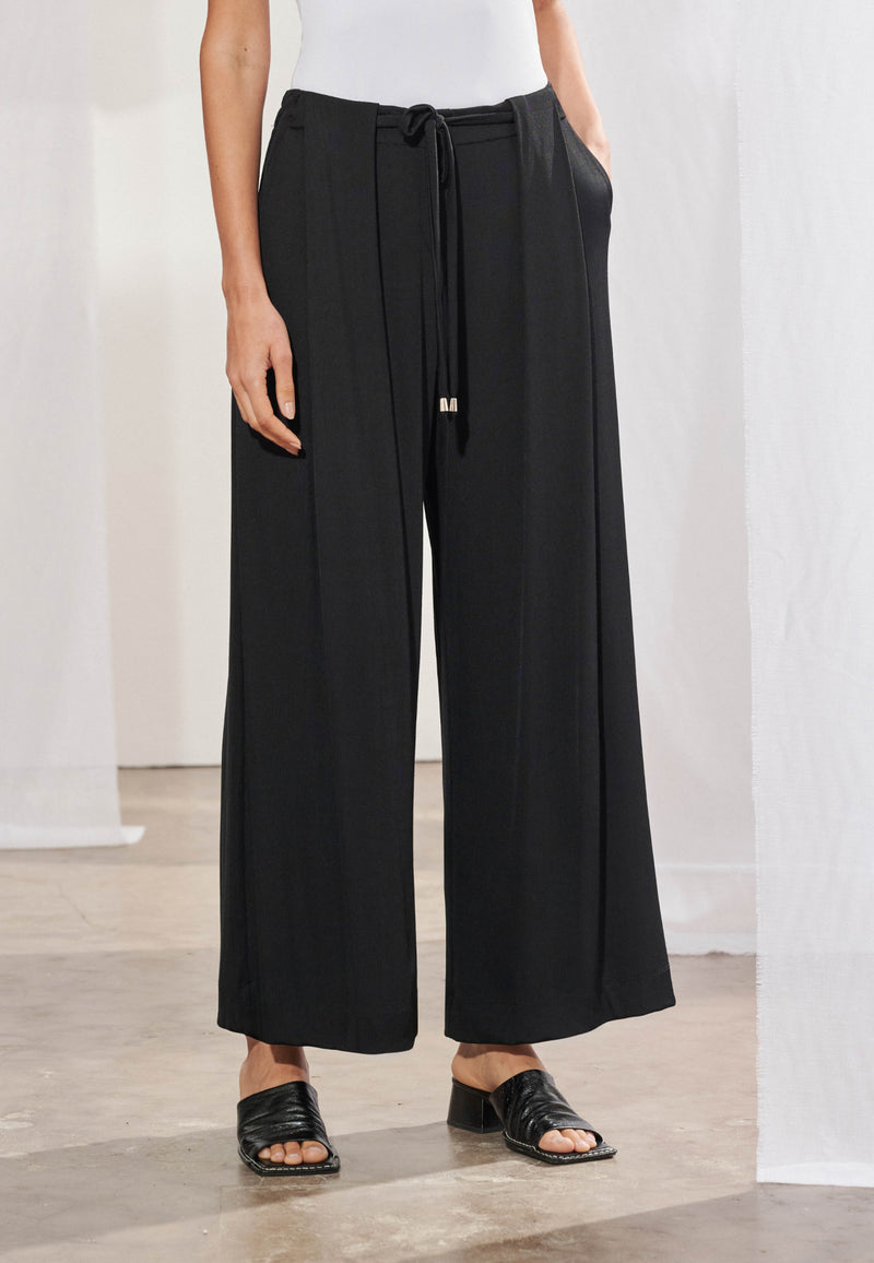 Calça pantalona longa em malha canelada, vista de frente. Modelagem ampla com pregas frontais profundas, cordão para amarração no cós com ponteiras metálicas e botão decorativo. Pernas largas com caimento fluido até o tornozelo.