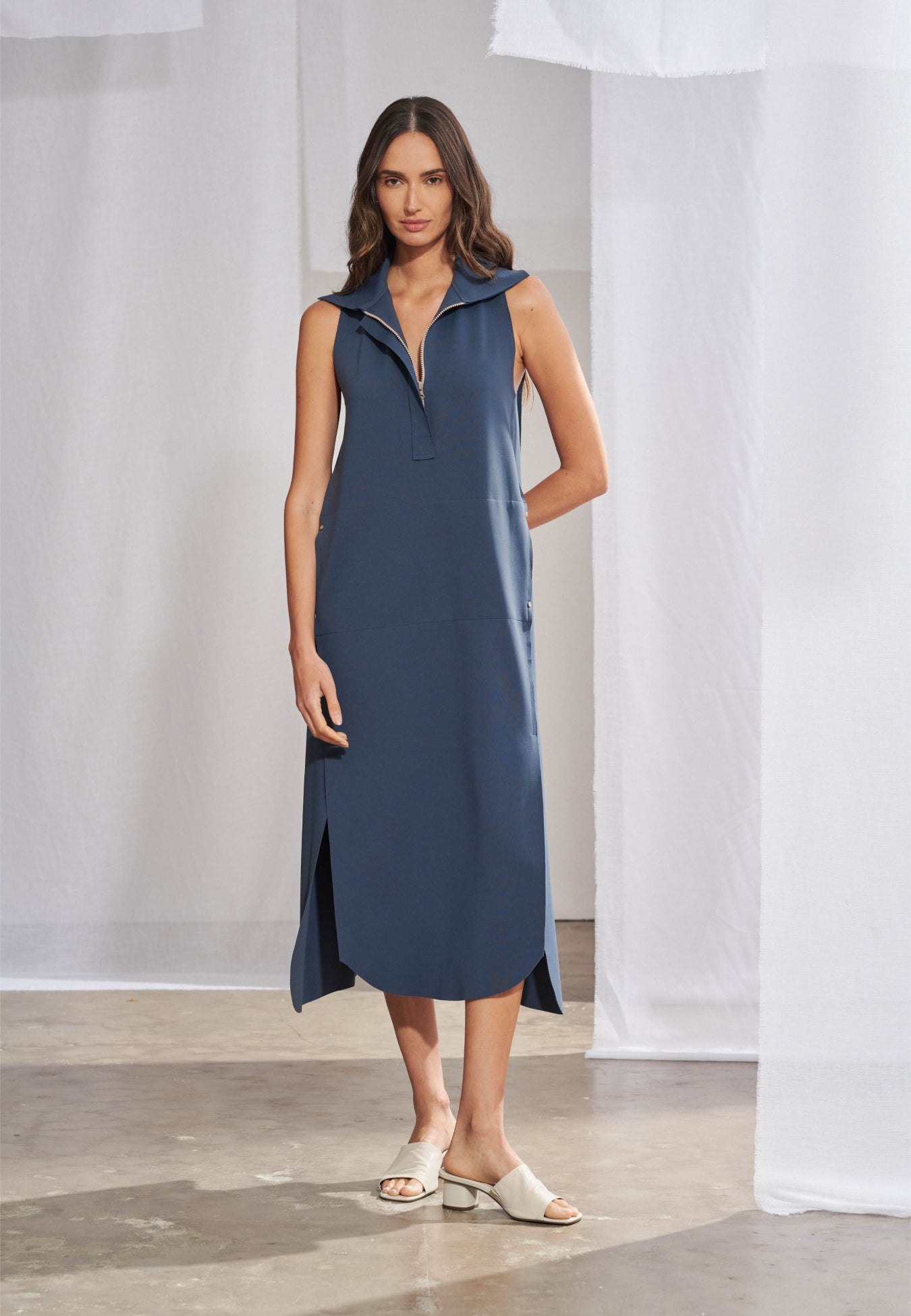 Vestido midi sem mangas de modelagem reta e caimento solto, em malha de alta densidade. Gola esporte estruturada com zíper frontal no decote, recortes verticais pespontados, fendas laterais com barra arredondada e bolsos embutidos nas costuras.