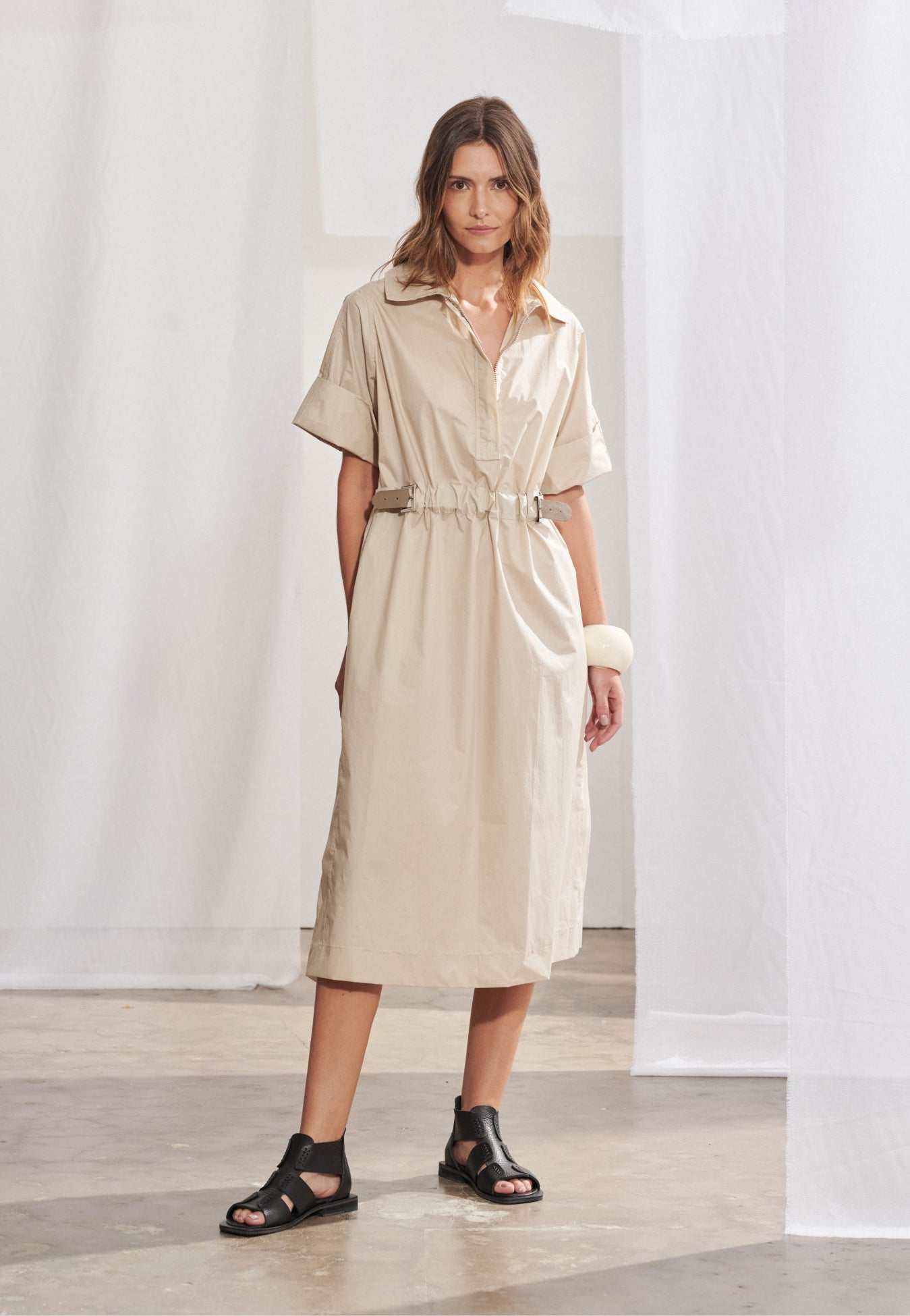 Vestido midi feminino de modelagem reta solta com mangas curtas de punho largo. Possui gola esporte, decote V com zíper tratorado aparente e ajuste na cintura regulável por cinto contrastante e fivela metálica. Saia de comprimento midi com fendas laterais e bolsos faca embutidos.