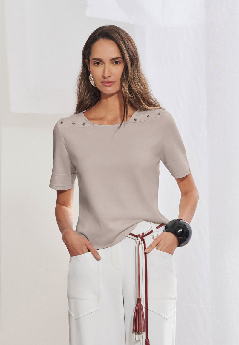 Blusa feminina de manga curta e modelagem reta. Possui decote redondo e recorte horizontal nos ombros com aplicação de quatro rebites metálicos em cada lado. As laterais se deslocam para as costas com acabamento geométrico que se repete na frente. A barra é assimétrica, ligeiramente alongada nas costas.