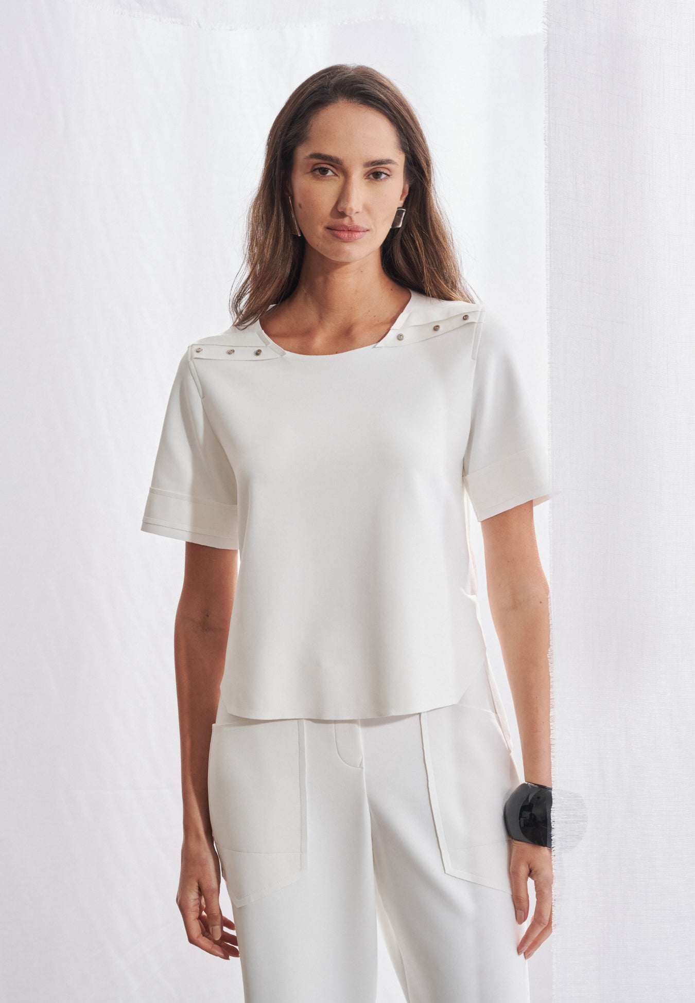 Blusa feminina de manga curta e modelagem reta. Possui decote redondo e recorte horizontal nos ombros com aplicação de quatro rebites metálicos em cada lado. As laterais se deslocam para as costas com acabamento geométrico que se repete na frente. A barra é assimétrica, ligeiramente alongada nas costas.