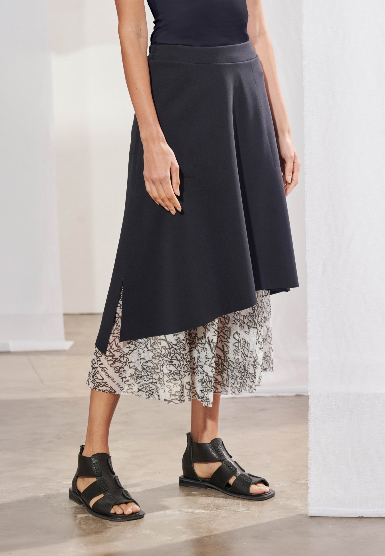 Saia midi feminina com modelagem evasê e sobreposição de duas camadas. A camada superior é feita em malha Tech, barra assimétrica diagonal e fendas laterais que revelam bolsos embutidos. A camada inferior é feita em tule fluido com estampa exclusiva em tons neutros, com comprimento que termina na altura da panturrilha.