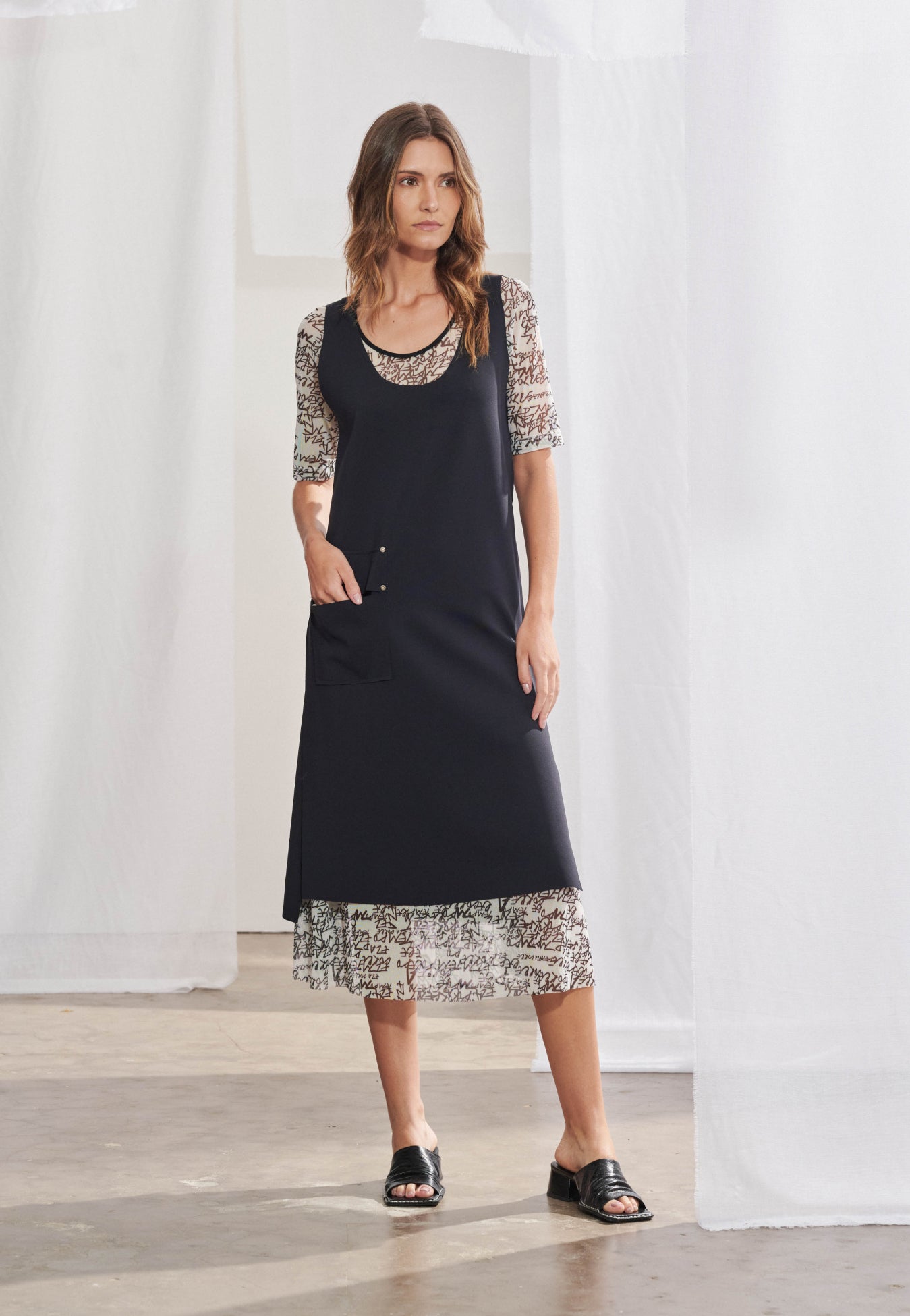 Vestido feminino midi de alfaiataria contemporânea em duas camadas. A base é uma blusa-vestido estampada com caligrafia preta e mangas curtas, sobreposta por um vestido preto de corte reto, com fendas laterais, bolso utilitário frontal com rebites metálicos e lapelas decorativas na parte posterior.