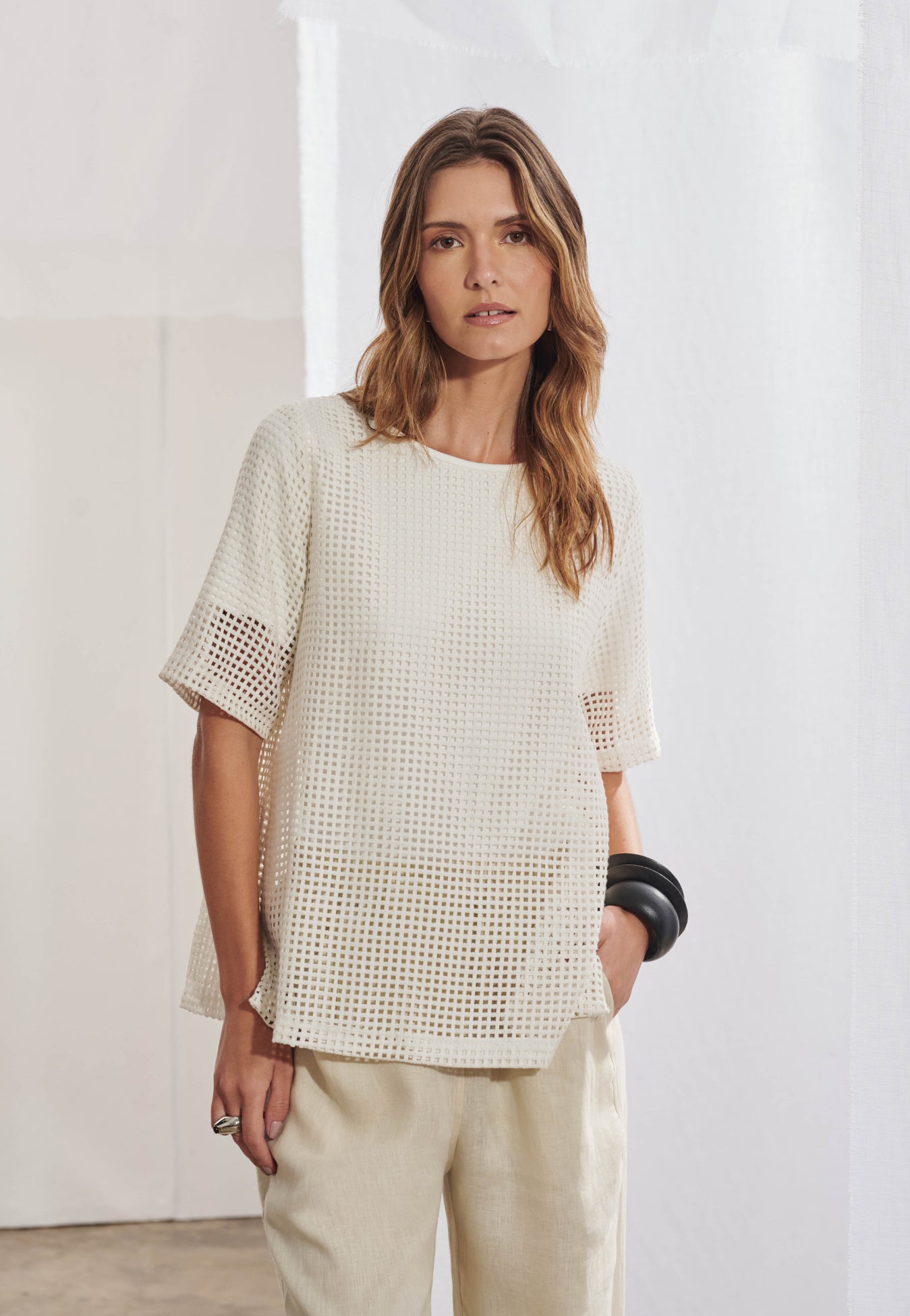 Blusa feminina ampla em malha telada com decote canoa acabado em viés. Mangas curtas com transparência visível na trama. Fendas laterais proeminentes na barra. Vista frontal mostrando a textura da tela no corpo e mangas.