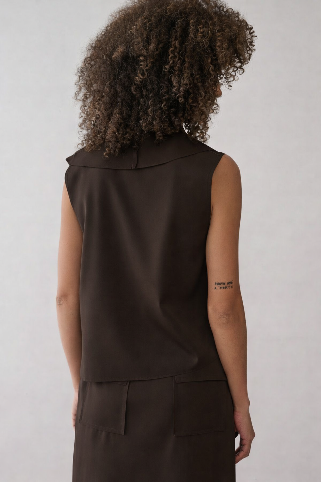 Modelo de costas veste uma blusa marrom sem mangas com gola aparente, combinada com saia reta da mesma cor com bolsos amplos.