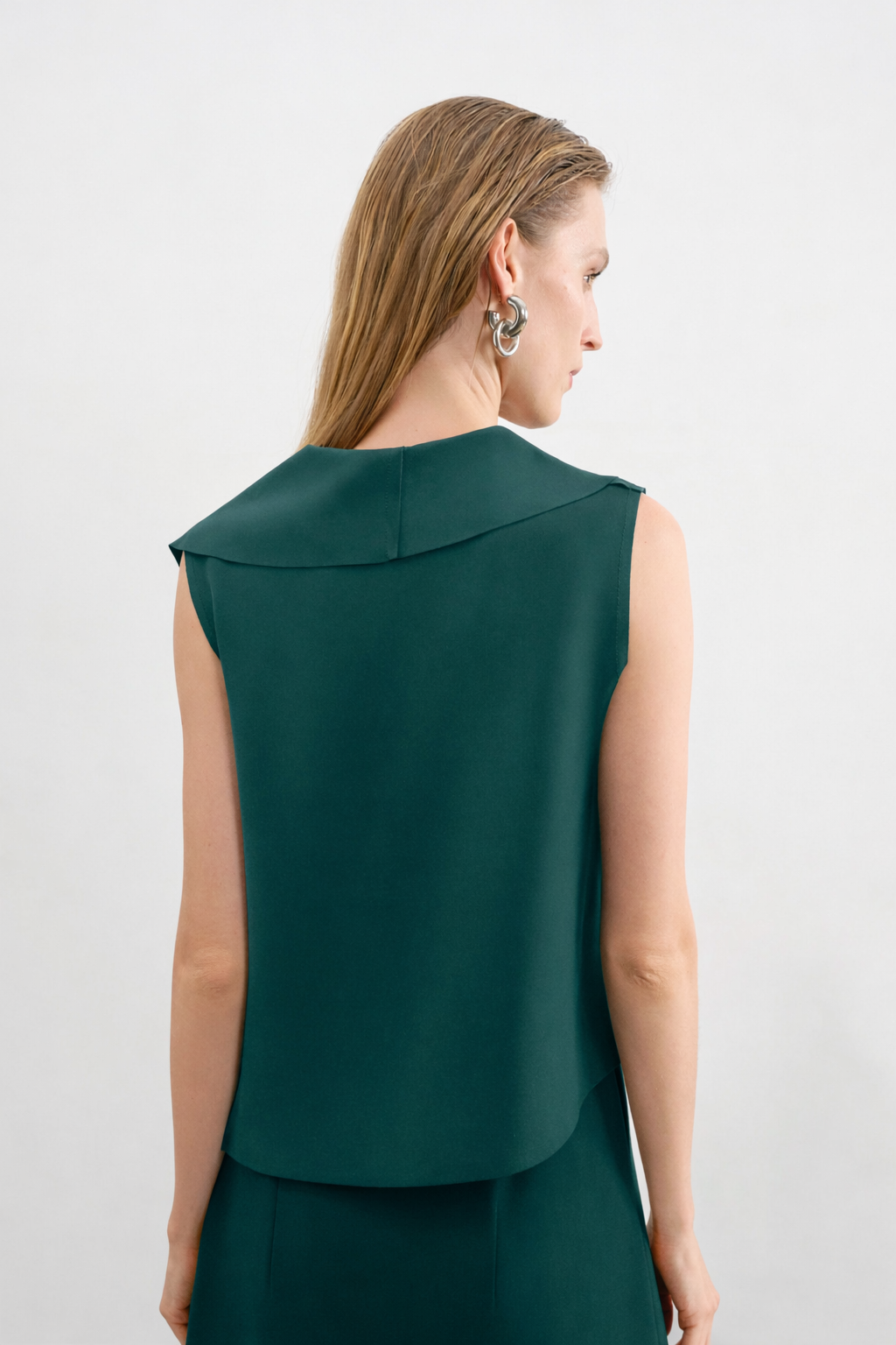 Modelo de costas veste uma blusa verde petróleo sem mangas com gola aparente, combinada com saia reta da mesma cor.