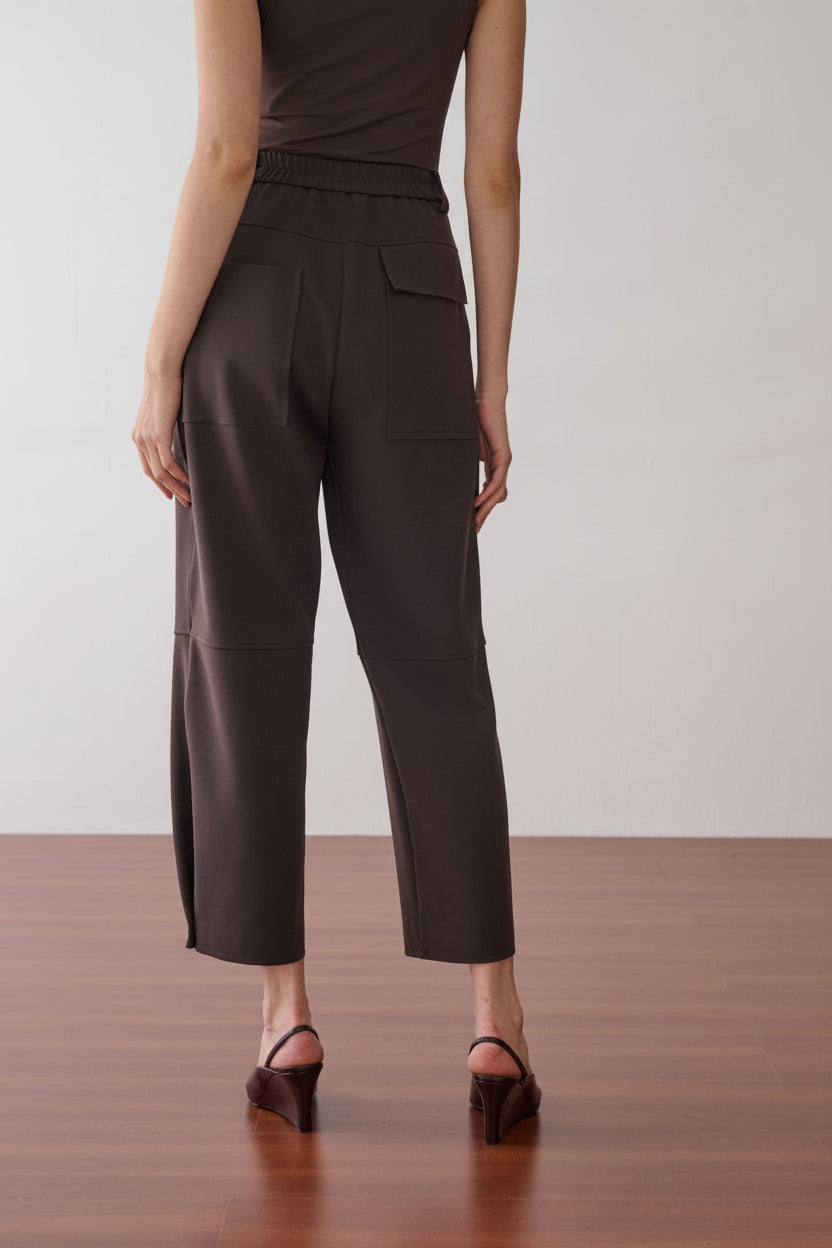 Modelo de costas veste uma calça marrom de cintura alta, modelagem reta, cós elástico e bolsos amplos.