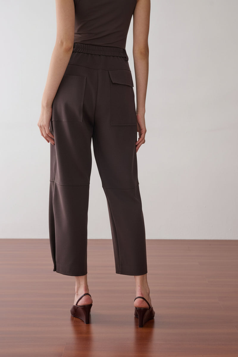 Modelo de costas veste uma calça marrom de cintura alta, modelagem reta, cós elástico e bolsos amplos.