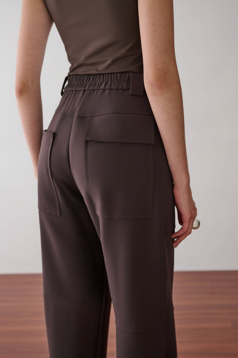 Modelo de costas veste uma calça marrom de cintura alta, modelagem reta, cós elástico e bolsos amplos.