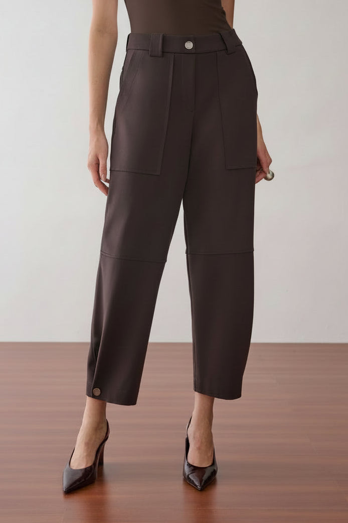 Modelo veste uma calça marrom de cintura alta e modelagem reta com bolsos frontais amplos, detalhe de botões nos punhos da barra e sapatos de bico fino.