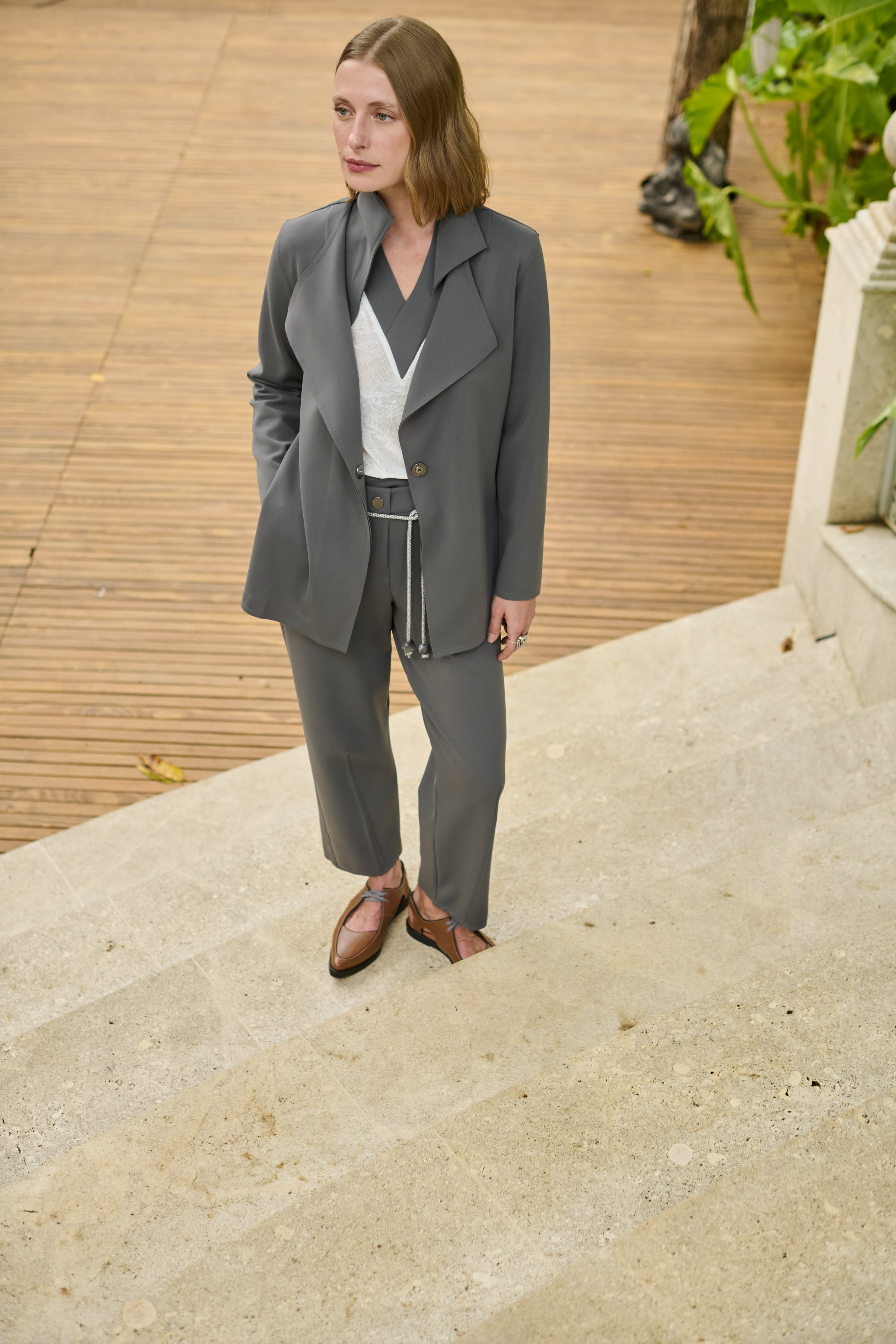 Modelo veste um conjunto cinza composto por blazer alongado de lapelas amplas e calça de alfaiataria, acompanhado de blusa branca com decote em "V", cordão prateado na cintura e sapatos marrons de bico fino.